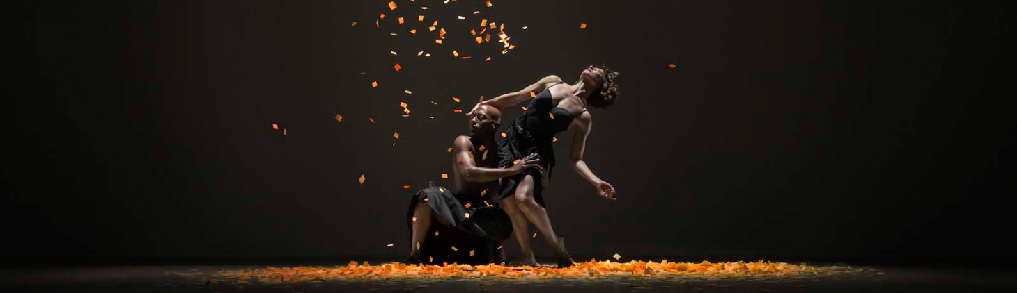 An image of two dancers, one crouched down next to the other dancer looking up towards the light while orange confetti falls